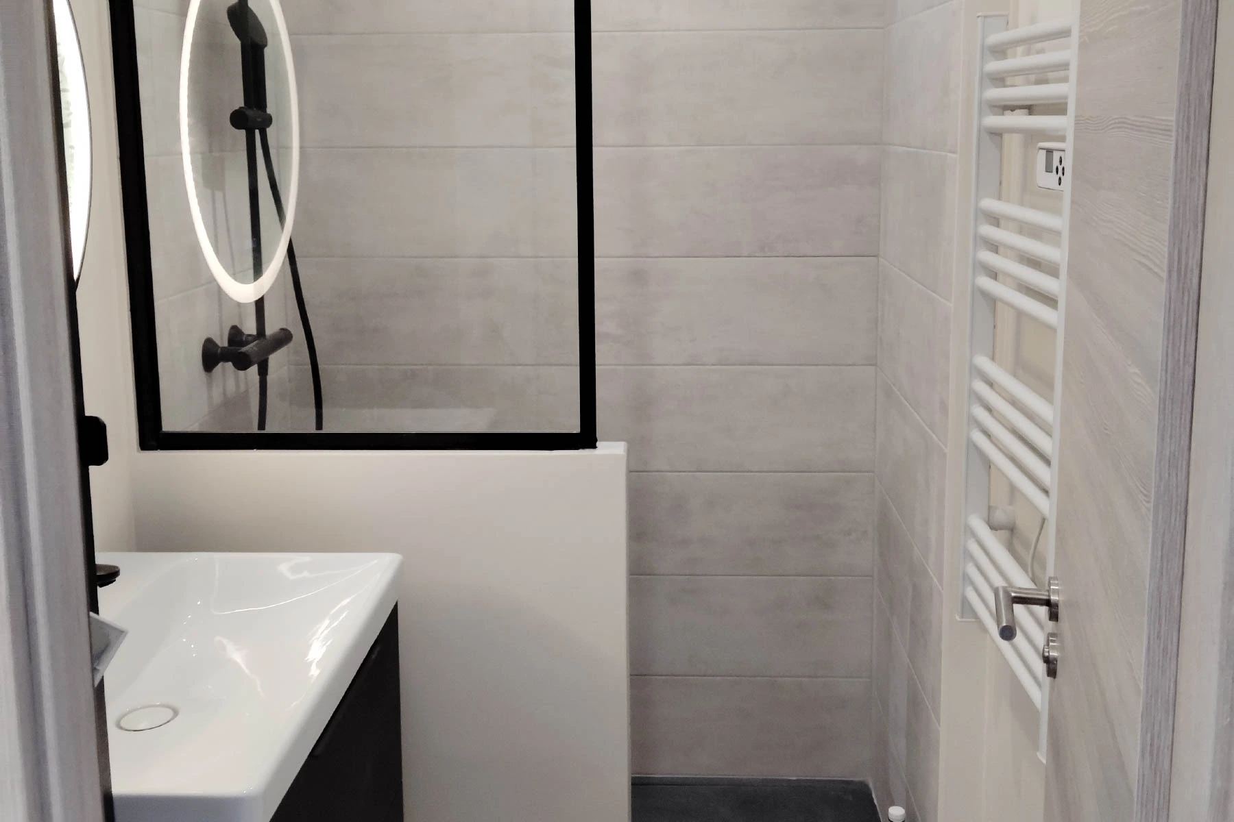 Salle de bain moderne entièrement rénovée à Nancy avec douche à l'italienne noire, miroir rond lumineux LED et carrelage effet béton gris